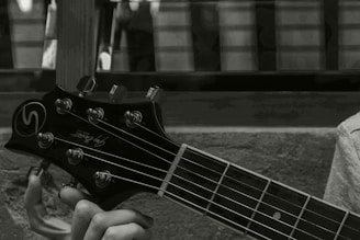 A close-up image of a person's hand holding the neck and headstock of a guitar. The guitar headstock has tuning pegs and a logo, with strings running down the neck. The background appears to be an out-of-focus window or wall.