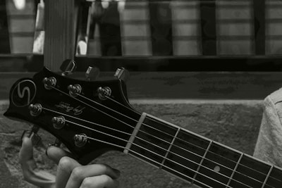 A close-up image of a person's hand holding the neck and headstock of a guitar. The guitar headstock has tuning pegs and a logo, with strings running down the neck. The background appears to be an out-of-focus window or wall.