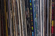 A collection of vinyl records is lined up vertically, with their spines displaying various album and artist names. The records appear tightly packed together, with a mix of modern and classic albums visible. The spines are colorful, indicating a diverse assortment of music genres.