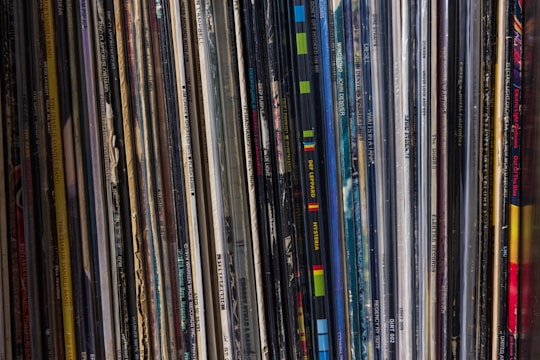 A collection of vinyl records is lined up vertically, with their spines displaying various album and artist names. The records appear tightly packed together, with a mix of modern and classic albums visible. The spines are colorful, indicating a diverse assortment of music genres.