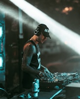 A DJ wearing headphones and a cap is focused on mixing music at a professional DJ controller. The background includes dim lighting and light beams creating a dynamic club atmosphere. The DJ has tattoos on his arms and appears engaged in his performance.