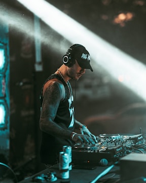A DJ wearing headphones and a cap is focused on mixing music at a professional DJ controller. The background includes dim lighting and light beams creating a dynamic club atmosphere. The DJ has tattoos on his arms and appears engaged in his performance.