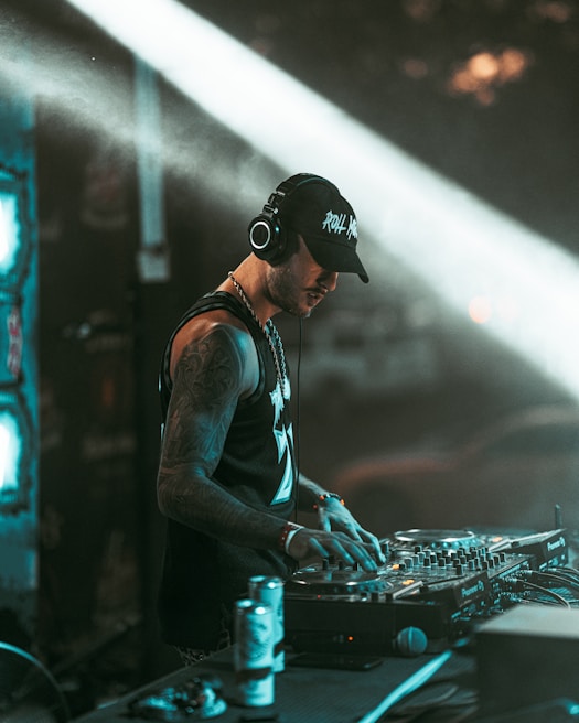 A DJ wearing headphones and a cap is focused on mixing music at a professional DJ controller. The background includes dim lighting and light beams creating a dynamic club atmosphere. The DJ has tattoos on his arms and appears engaged in his performance.