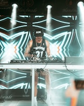 A DJ wearing a black cap and headphones stands behind a mixing console performing at an event. The DJ is dressed in a sleeveless sports jersey and leopard print shorts, with bright stage lights illuminating the scene. The background features a vibrant LED screen with abstract, geometric patterns in shades of blue and white.
