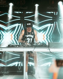 A DJ wearing a black cap and headphones stands behind a mixing console performing at an event. The DJ is dressed in a sleeveless sports jersey and leopard print shorts, with bright stage lights illuminating the scene. The background features a vibrant LED screen with abstract, geometric patterns in shades of blue and white.