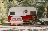 A vintage trailer with a white and red color scheme is parked on a sunny driveway. In front of it, a white wicker bench with two red pillows featuring snowflake designs is placed on a small rug. A holiday wreath hangs on the trailer door, and a red wagon nearby holds a small, decorated Christmas tree. To the right is a vintage sports car partially visible under lush greenery.