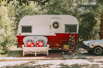 A vintage trailer with a white and red color scheme is parked on a sunny driveway. In front of it, a white wicker bench with two red pillows featuring snowflake designs is placed on a small rug. A holiday wreath hangs on the trailer door, and a red wagon nearby holds a small, decorated Christmas tree. To the right is a vintage sports car partially visible under lush greenery.