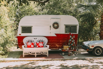 A vintage trailer with a white and red color scheme is parked on a sunny driveway. In front of it, a white wicker bench with two red pillows featuring snowflake designs is placed on a small rug. A holiday wreath hangs on the trailer door, and a red wagon nearby holds a small, decorated Christmas tree. To the right is a vintage sports car partially visible under lush greenery.