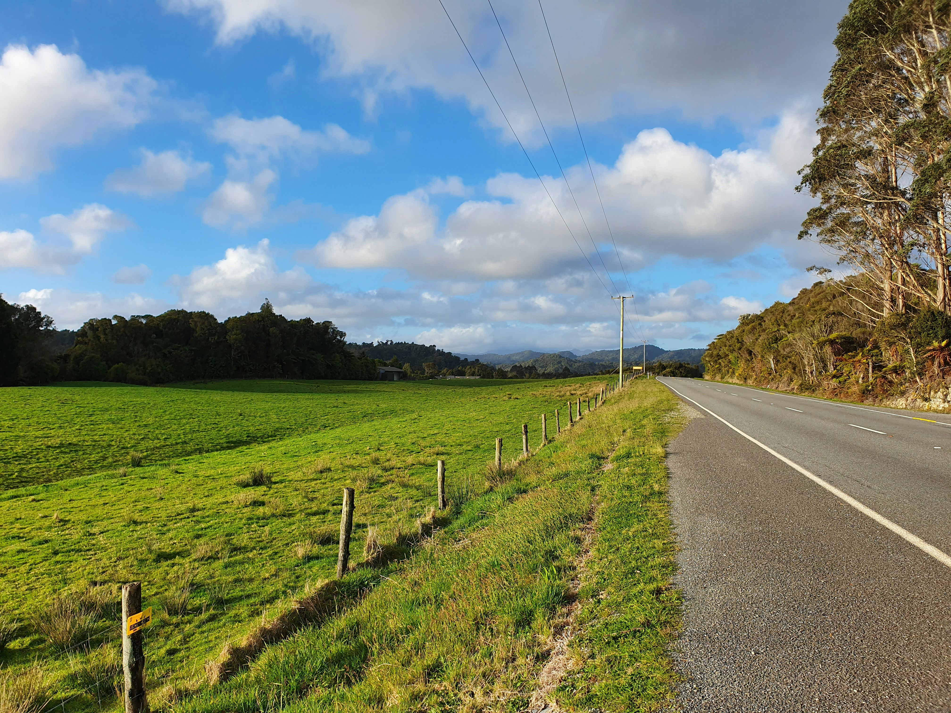 gray concrete road between green grass field under blue sky and white clouds during daytime
