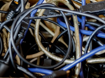 A tangled jumble of various cables and wires, including black, blue, and transparent ones, appear intertwined in a chaotic manner. Connectors and plugs are visible at the ends of some wires, with different textures and thicknesses of the cables contributing to the visual complexity.