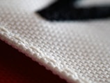 A close-up view of a textured fabric material, showcasing the intricate weave of the threads. The white cloth appears to be craft or industrial-grade with a clear focus on its structure, highlighting each individual fiber and strand.