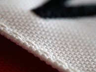 A close-up view of a textured fabric material, showcasing the intricate weave of the threads. The white cloth appears to be craft or industrial-grade with a clear focus on its structure, highlighting each individual fiber and strand.