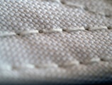 A close-up view of a textured fabric surface, showing intricate stitching and woven threads. The fabric appears to be tightly knit, with visible threads that form a repetitive pattern.