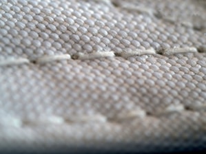 A close-up view of a textured fabric surface, showing intricate stitching and woven threads. The fabric appears to be tightly knit, with visible threads that form a repetitive pattern.