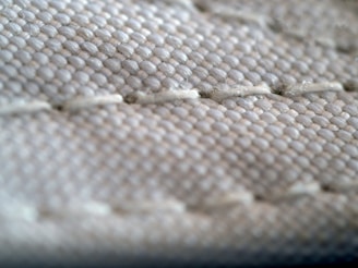 A close-up view of a textured fabric surface, showing intricate stitching and woven threads. The fabric appears to be tightly knit, with visible threads that form a repetitive pattern.