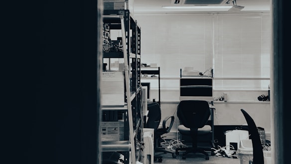 An office or workspace with two chairs around a table, a shelving unit filled with books and files on one side, several computer monitors, and various cables visible on the ground. The room is lit by natural light coming through partially closed blinds.