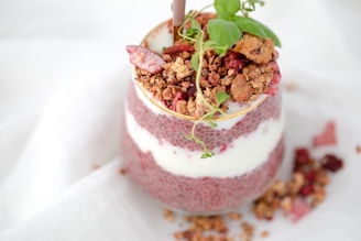 A layered dessert in a glass consisting of pink chia seed pudding and white yogurt, topped with granola, red berries, and fresh green leaves. Some granola and berries are scattered around the base.