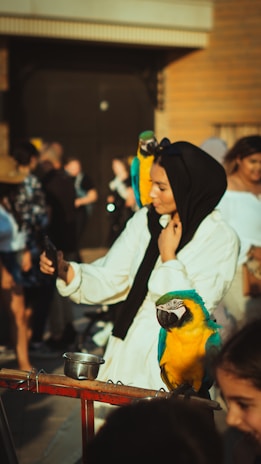 A woman wearing a black hijab and white dress is taking a selfie in an outdoor setting. She has a colorful parrot perched on her shoulder and another one on a stand in front of her. The background is blurred with several people and a large wooden door visible in the distance.