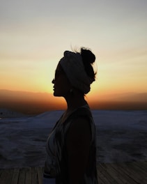 A silhouette of a woman with a headscarf stands against a vibrant sunset sky, which casts a warm glow over an expansive, open landscape.