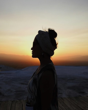 A silhouette of a woman with a headscarf stands against a vibrant sunset sky, which casts a warm glow over an expansive, open landscape.