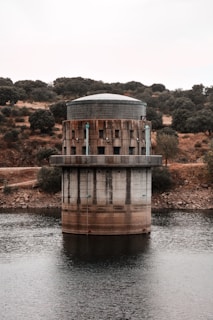 A cylindrical structure with a flat, dome-like top positioned in the middle of a water body. It features a rugged, industrial design with visible pipes and dark metal elements. Surrounding the water are rocky, dry landscapes dotted with sparse trees and bushes, under an overcast sky.