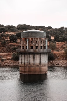 A cylindrical structure with a flat, dome-like top positioned in the middle of a water body. It features a rugged, industrial design with visible pipes and dark metal elements. Surrounding the water are rocky, dry landscapes dotted with sparse trees and bushes, under an overcast sky.