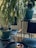 An indoor setting with several potted plants on metal stands near a window. Two chairs with wire frames and wooden armrests are situated beside the plants. Smoke, possibly from incense, rises gently from an earthen pot on a black tray table.