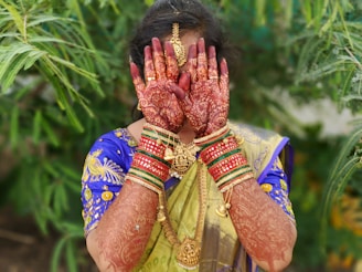 A person dressed in traditional attire is covering their face with intricately hennaed hands. The outfit is colorful, featuring a blend of blue and gold, and adorned with ornate gold jewelry. The background is lush with green foliage, enhancing the vibrant scene.