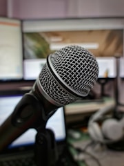 A close-up view of a professional microphone with a metallic mesh head, positioned in front of computer screens and headphones in the background. The setting suggests a workspace or studio environment for recording or broadcasting.