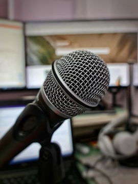 A close-up view of a professional microphone with a metallic mesh head, positioned in front of computer screens and headphones in the background. The setting suggests a workspace or studio environment for recording or broadcasting.