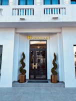 A modern entrance with a black glass door framed by two meticulously shaped topiary plants in bronze pots. Above the door, a golden sign reading 'H Victoriei 139' is prominently displayed. The façade is white with decorative elements and blue-tinted windows.