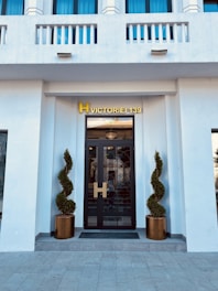 A modern entrance with a black glass door framed by two meticulously shaped topiary plants in bronze pots. Above the door, a golden sign reading 'H Victoriei 139' is prominently displayed. The façade is white with decorative elements and blue-tinted windows.