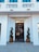 A modern entrance with a black glass door framed by two meticulously shaped topiary plants in bronze pots. Above the door, a golden sign reading 'H Victoriei 139' is prominently displayed. The façade is white with decorative elements and blue-tinted windows.