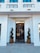 A modern entrance with a black glass door framed by two meticulously shaped topiary plants in bronze pots. Above the door, a golden sign reading 'H Victoriei 139' is prominently displayed. The façade is white with decorative elements and blue-tinted windows.