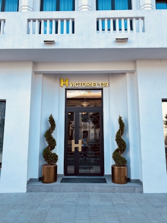 A modern entrance with a black glass door framed by two meticulously shaped topiary plants in bronze pots. Above the door, a golden sign reading 'H Victoriei 139' is prominently displayed. The fa&ccedil;ade is white with decorative elements and blue-tinted windows.