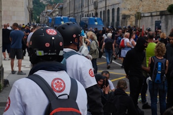 A crowded street scene with individuals wearing helmets and medical insignia, possibly medical personnel, amidst a large group of people. Blue police vehicles are visible in the background. The atmosphere appears busy and slightly tense.