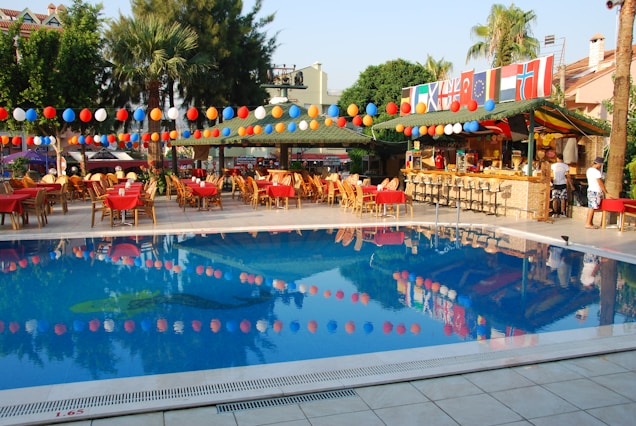 An outdoor area with a swimming pool surrounded by tables and chairs covered with red tablecloths. Colorful balloons are strung above, and a bar area with flags from different countries is visible. There are palm trees and greenery providing a tropical atmosphere.