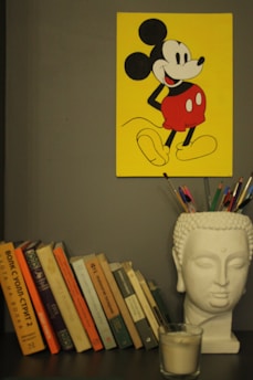 A collection of books is arranged on a shelf beside a white Buddha head sculpture, which holds colored pencils. Above the shelf, there is a bright yellow poster featuring a cartoon character in red shorts.