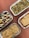 Four foil containers filled with different kinds of pasta dishes are arranged on a reddish-brown surface. The top right container contains fettuccine with mushrooms and herbs. Below it, a pasta dish topped with melted cheese and herbs. The bottom left container holds a creamy lasagna, while the top left features pasta with meat, mayonnaise, slices of lemon, and red chili slices.