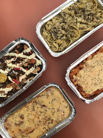 Four foil containers filled with different kinds of pasta dishes are arranged on a reddish-brown surface. The top right container contains fettuccine with mushrooms and herbs. Below it, a pasta dish topped with melted cheese and herbs. The bottom left container holds a creamy lasagna, while the top left features pasta with meat, mayonnaise, slices of lemon, and red chili slices.