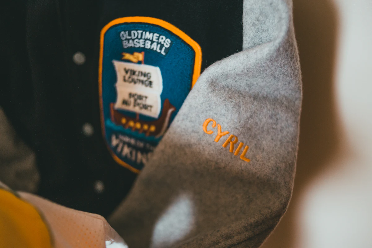 Patch bordir berbentuk perisai dijahit keliling pada dada jaket varsity, dengan nama bordir pada lengan wool; tekstur benangnya tebal dan pinggiran patchnya dijahit rapi.