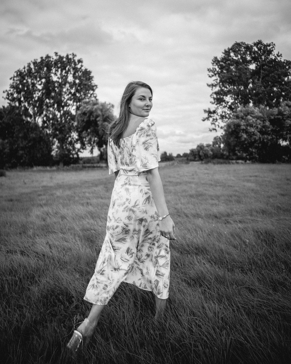 Foto en escala de grises de la muchacha con vestido floral de pie en el campo de hierba