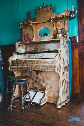 A vintage piano with ornate carvings is positioned against a teal wall. The piano is accented with intricate detailing and a small oval mirror at the top center. There are decorative items such as a brass pitcher and an antique book resting on the piano's shelf. The scene includes a rustic wooden stool with a circular seat in front of the piano.