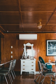 A rustic room features a white dresser with ornate detailing and a mirror placed against a wooden paneled wall. On top of the dresser, there are metal teapots and a framed photo. The room is furnished with silver chairs surrounding tables, and has a wooden floor and ceiling with a small chandelier. An air conditioning unit is mounted above the dresser, and a framed artwork depicting a winter scene hangs on the wall.