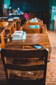 A classroom with wooden desks and chairs, featuring textbooks stacked neatly on the surfaces. The focus is on the empty desks, while a teacher and a student can be seen in the blurred background near a chalkboard filled with writing.