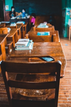 A classroom with wooden desks and chairs, featuring textbooks stacked neatly on the surfaces. The focus is on the empty desks, while a teacher and a student can be seen in the blurred background near a chalkboard filled with writing.