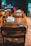 A classroom with wooden desks and chairs, featuring textbooks stacked neatly on the surfaces. The focus is on the empty desks, while a teacher and a student can be seen in the blurred background near a chalkboard filled with writing.