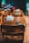 A classroom with wooden desks and chairs, featuring textbooks stacked neatly on the surfaces. The focus is on the empty desks, while a teacher and a student can be seen in the blurred background near a chalkboard filled with writing.