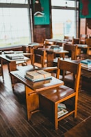 A classroom with wooden desks and chairs arranged in neat rows, each desk topped with stacks of books. The room features large windows that let in natural light, creating a warm and nostalgic atmosphere. The floor is made of polished wood, and the walls are decorated with a dark turquoise color.
