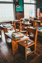 A classroom with wooden desks and chairs arranged in neat rows, each desk topped with stacks of books. The room features large windows that let in natural light, creating a warm and nostalgic atmosphere. The floor is made of polished wood, and the walls are decorated with a dark turquoise color.
